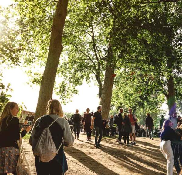 På en grussti under flere træer går en masse mennesker op imod de plastiktelte der står i baggrunden.