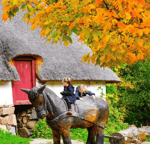 Børn leger på en hest udskåret af træ foran et gammelt stråtækt hus og et træ med gul-orange blade på