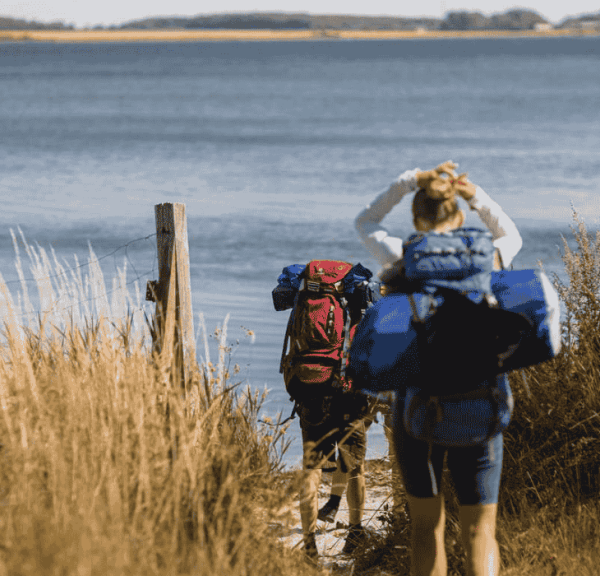 To vandrere er på vej igennem højt græs ned til havet, og de bærer begge vandreudstyr og rygsæk
