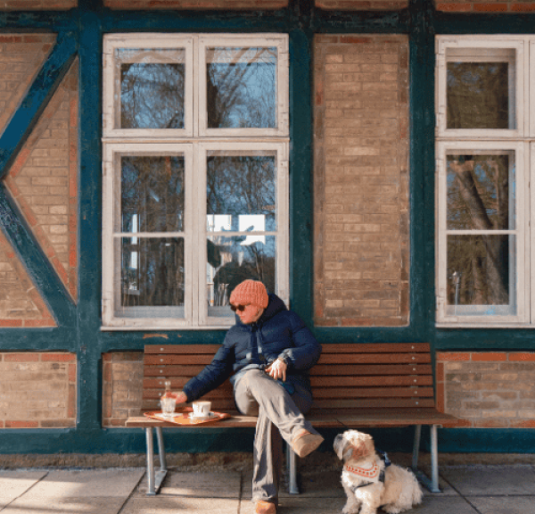 En kvinde iført vintertøj sidder ude foran en kaffebar med sin hund i snor og kaffe på en bakke