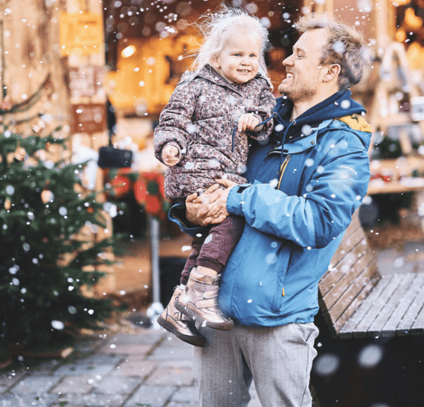 En mand holder et smilende barn i sine arme, mens han står udenfor i sneen, tæt på et juletræ og festlige julemarkedsboder.