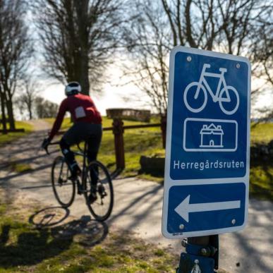 i forgrunden er et blåt skilt med en cykel, pil til venstre og tekst hvor der står Herregårdsruten. I baggrunden cykler en mand på racercykel ned af en sti der går mellem træer. 