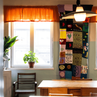 Et rum med farverige gardiner om et lysindrammet vindue og et sammenstrikket tæppe der hænger på væggen ved siden af