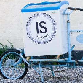 Lyseblå is-cykel med logoet "Is fra Skarø" står parkeret ved en væg.