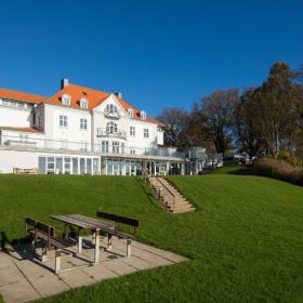 Stort, hvidt hotel med rødt tag beliggende i en stor have. Bænkesæt i forgrunden.