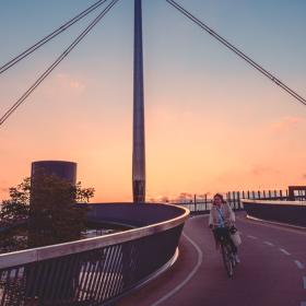 Byens bro i Odense vist i en flot solnedgang med gule og orange nuancer. En cyklist kommer cyklende på vej ned af broen.
