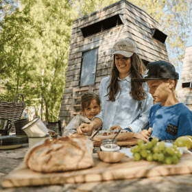 En familie bestående af en mor, en datter og en søn, iført sommertøj, laver mad udendørs foran et shelter