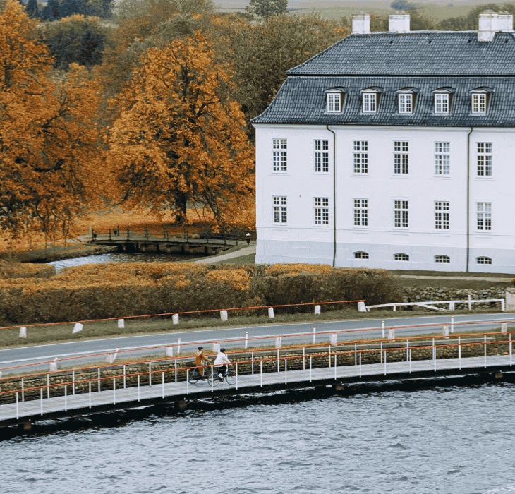 To cyklister kører på en cykelsti ude på vandet foran et hvidt gods en overskyet dag