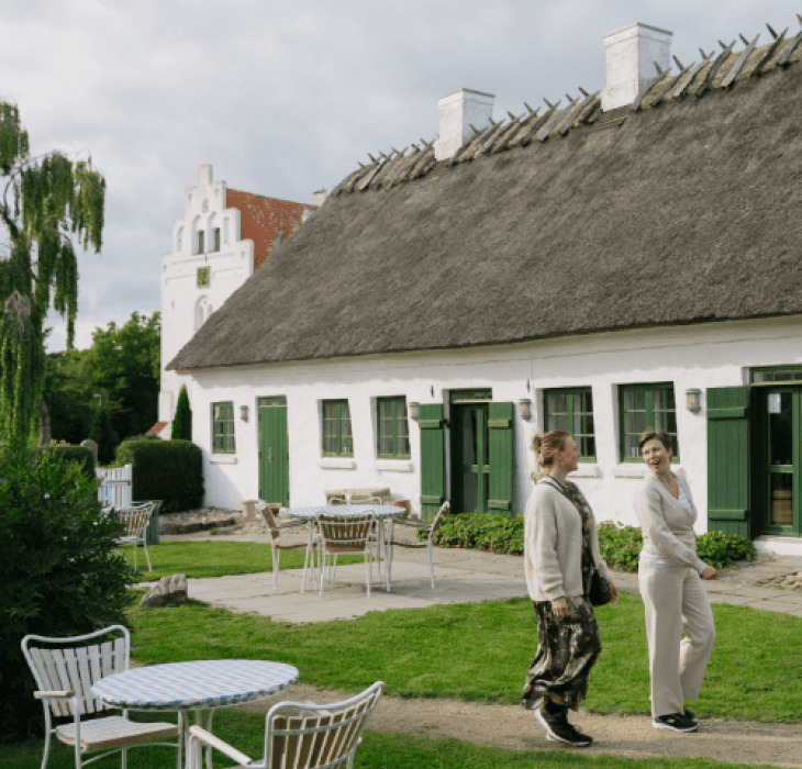 To kvinder går foran en gammel kirkelade med stråtækt tag og en hvid kirke i baggrunden