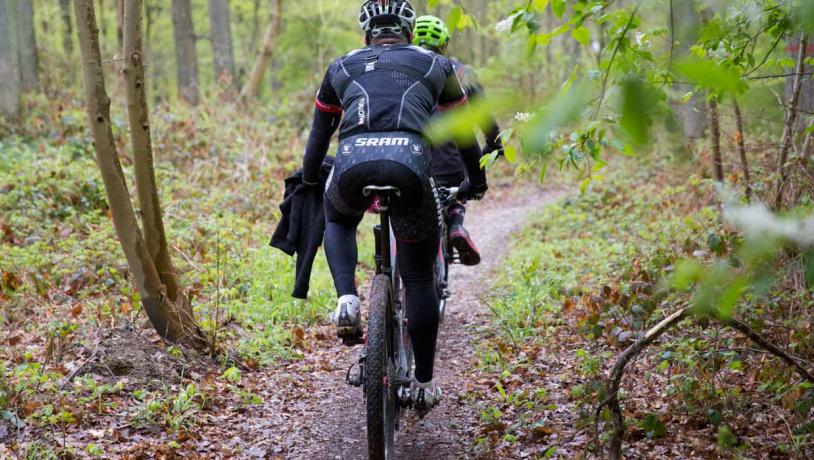 To mountainbikere cykler i en skov, omringet af træer og vildt mos.