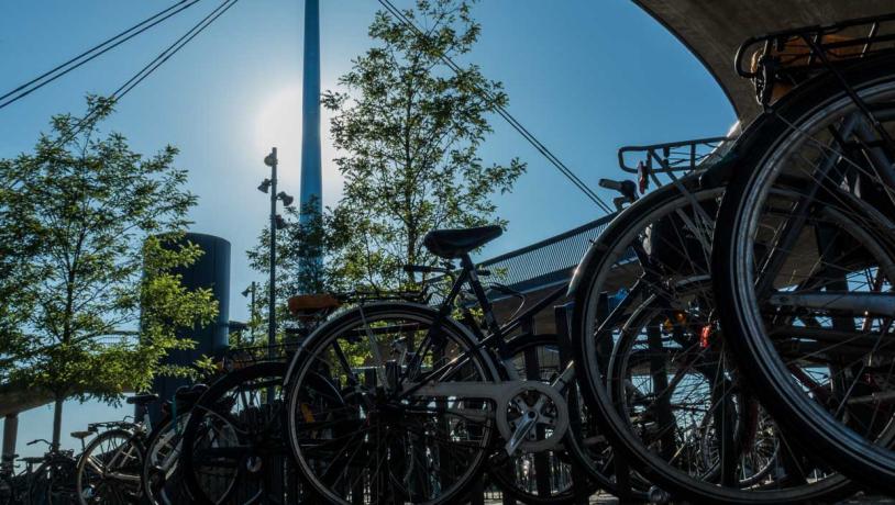 Parkerede cykler ved Byens Bro i Odense. Broen rejser sig mod en blå himmel og en bagende sol i baggrunden.