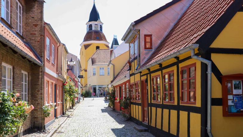 En malerisk gade i Faaborg med brostensbelægning og farverige huse med bindingsværk. Husene har levende gule og røde facader med traditionelle røde tegltage. I baggrunden tårner Faaborgs karakteristiske klokketårn sig op med sin røde kuppel og spir, hvilket tilføjer et charmerende historisk element til scenen. Det er en solrig dag med blå himmel og stokroser i fuldt flor, der tilføjer liv til gadebilledet.