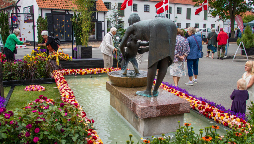 Billede af springvand med statue i midten, dekoreret med kant af roser i gule, orange og røde farver.