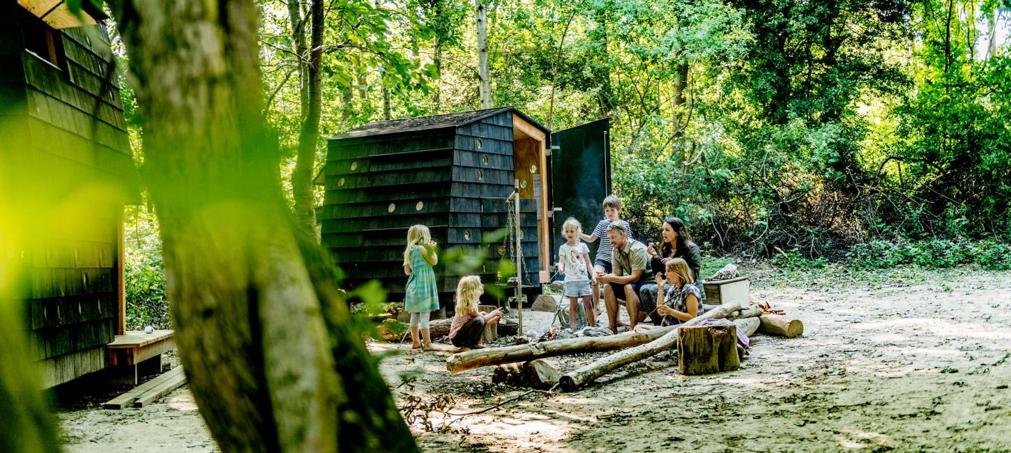 Fem børn og to voksne samlet om et bål i skoven. Bag ved dem ses et arkitekttegnet shelter.