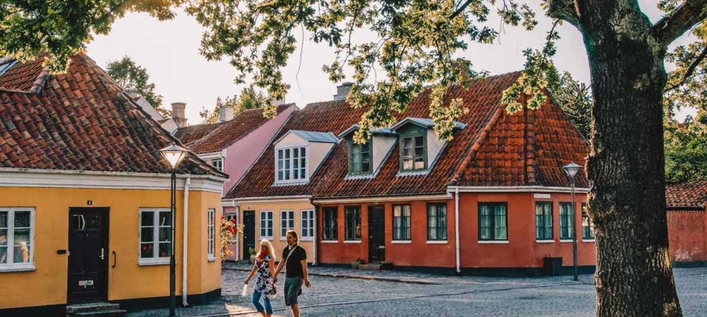 Et par går hånd i hånd på en brostensgade. I baggrundenden er fire bygninger i forskellige farver. Solen skinner gennem en trækrone. 