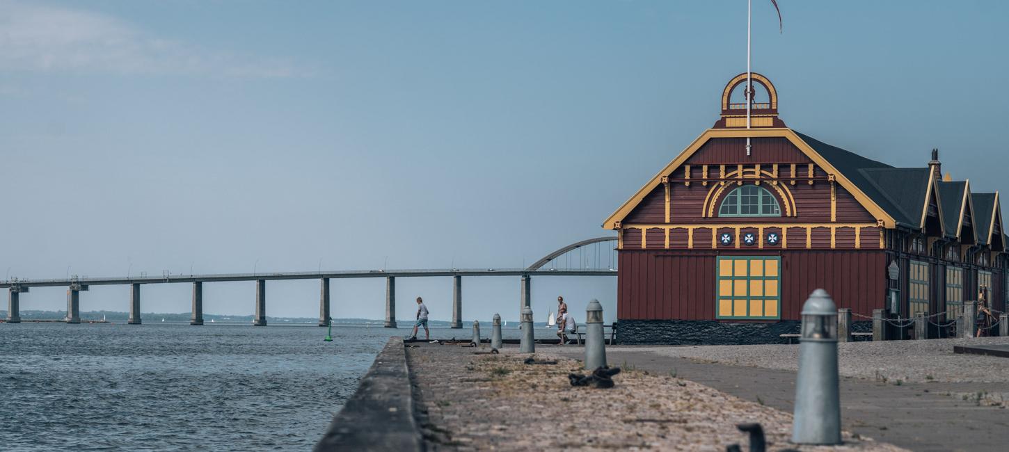 Ved en havn ses et pakhus i gult og rødt træ. I baggrunden kan man se Langelandsbroen.