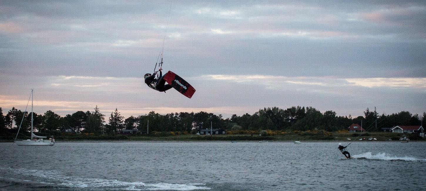 Midt i billedet flyver en kitesurfer over havet. Til venstre i billedet kan man se en sejlbåd på vej ud af billedet. Til højre er endnu en kitesurfer. Han surfer på vandet. I horisonten kan man se en skov på land. 