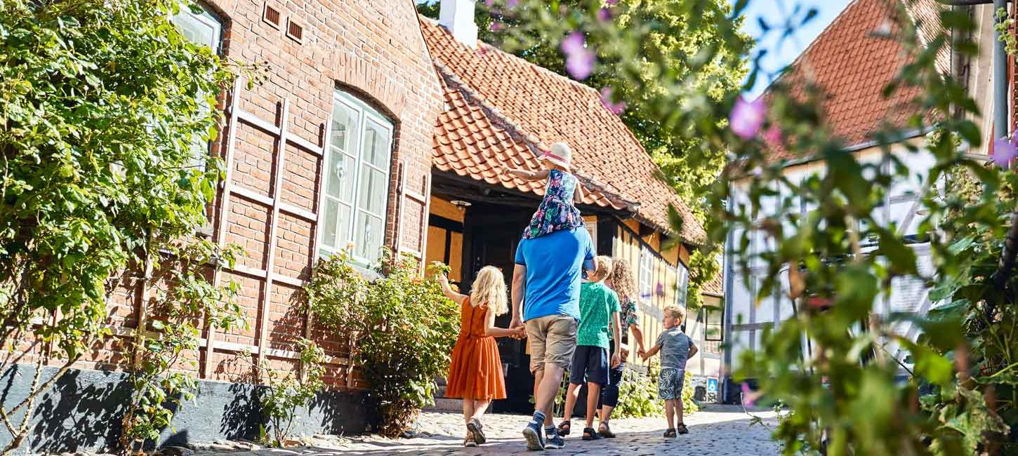 Familie med to voksne og tre børn går ned ad en brostensbelagt gade med bindingsværkshuse og stokroser.