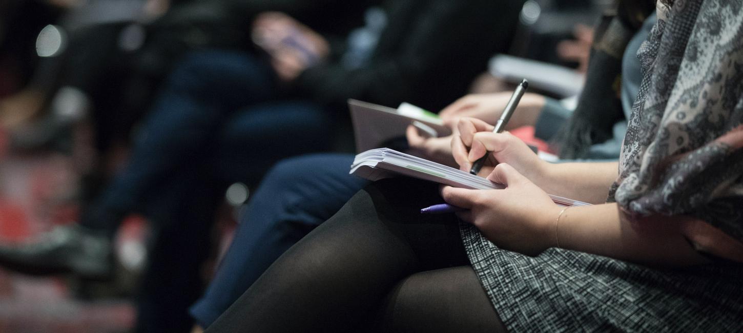 Et nærbilede af kvindehænder der skriver i en notes bog liggende på hendes krydsede ben. Man kan ikke se hendes hoved. I baggrunden kan man se at hun sidder på en række, hvor nogle mennesker gør det samme.