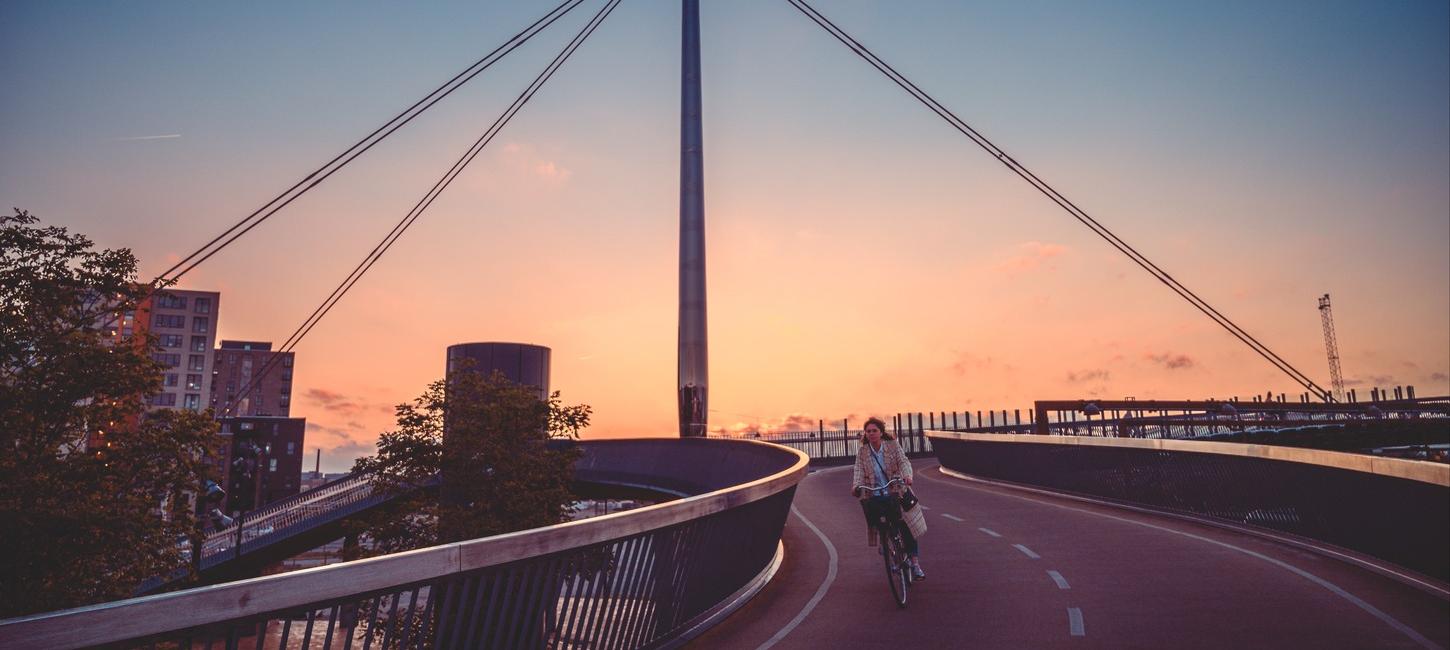 Byens bro i Odense vist i en flot solnedgang med gule og orange nuancer. En cyklist kommer cyklende på vej ned af broen.