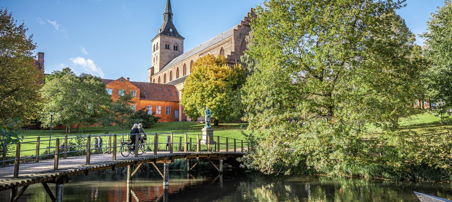 En idyllisk grøn park med en stor kirke med et spidst tårn i baggrunden. Kirken, der har en klassisk arkitektonisk stil, er omgivet af frodige træer i fuldt løv. Foran kirken er der en lille bro over en rolig å, hvor en person krydser på cykel. Himlen er klar og blå.