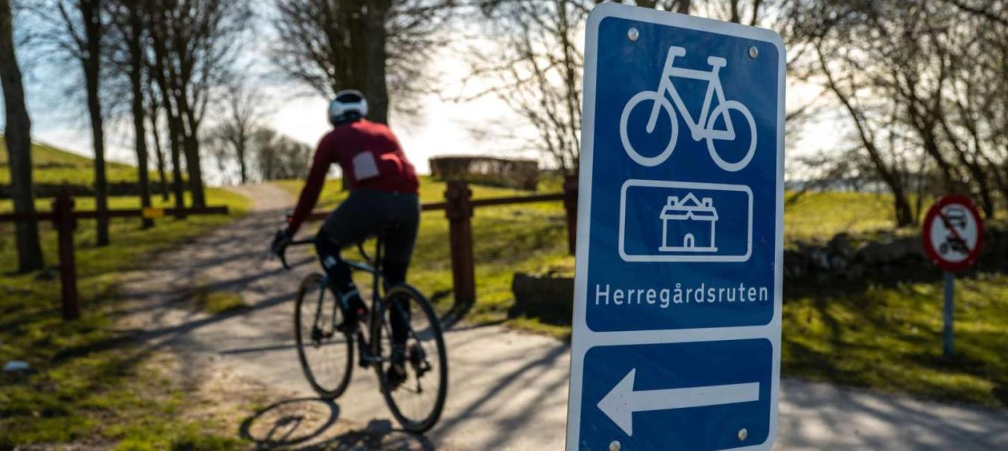 i forgrunden er et blåt skilt med en cykel, pil til venstre og tekst hvor der står Herregårdsruten. I baggrunden cykler en mand på racercykel ned af en sti der går mellem træer. 