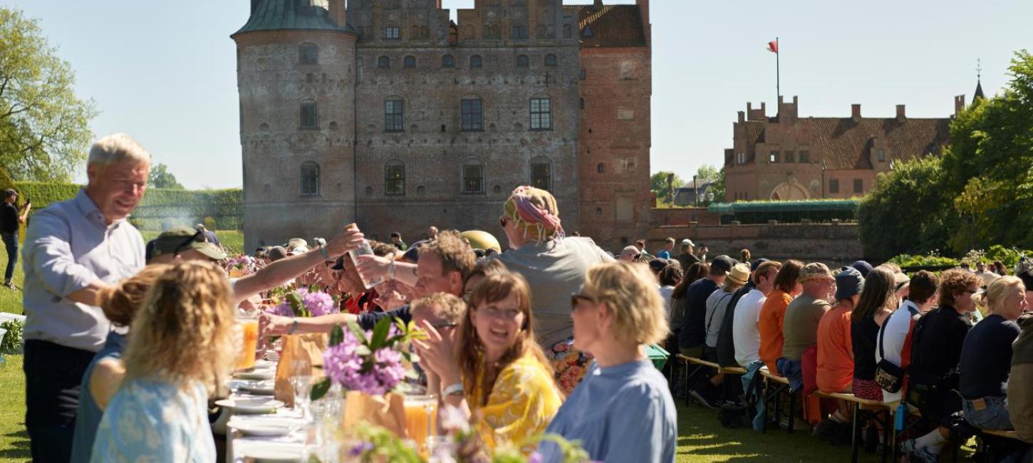 Mange mennesker sidder ved langborde under åben himmel og spiser foran Egeskov Slot.