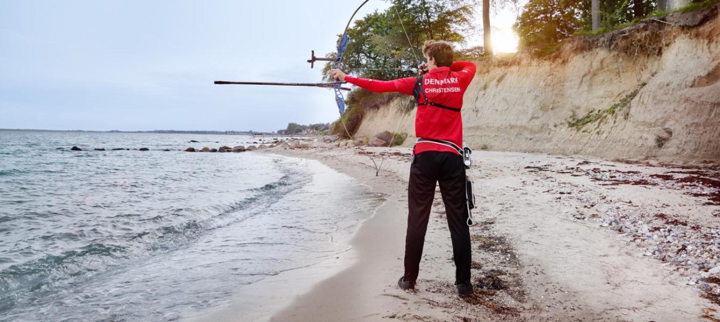 Bueskytte står på strand med ryggen til med buen spændt og sigter ud over vandet.