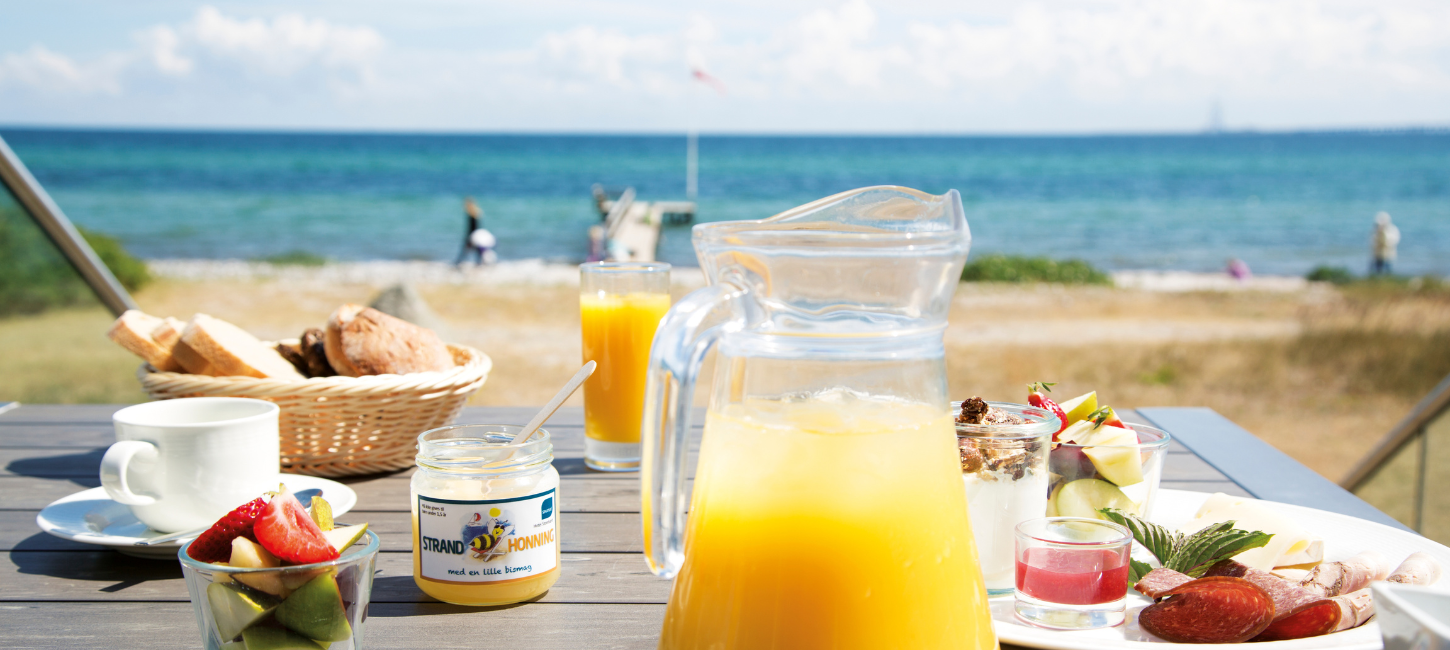 Juice, honning og andre lækre morgenmadsprodukter i forgrunden,. Strand, hav og blå himmel i baggrunden.