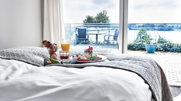 I fodenden af en dobbeltseng med hvid lagner står en bakke med morgenmad. Bagved sengen er der gennem vinduet udsigt til en terrasse der har mark og skov bag sig.
