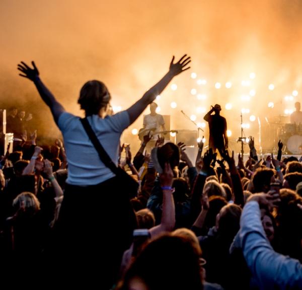 Foran en scene står et band oplyst af orange lys. Publikum står med begge hænder i vejret. En kvinde sidder på skulderen af en anden og hun har begge arme over hovedet. 