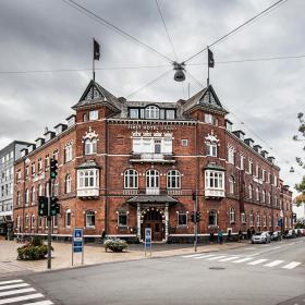 Stor, rød, symmetrisk, slotsagtig bygning i bymiljø med fodgængerfelter og lyssignaler i forgrunden under en mørk og faretruende himmel.