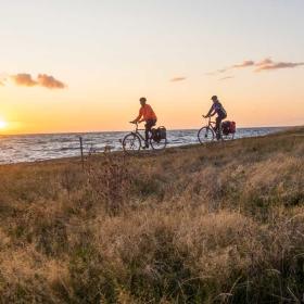 To cyklister kører langs med vandet med solnedgang i baggrunden og bølgende græs i forgrunden.