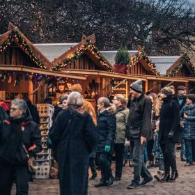 En brostensplads er fyldt med mennesker pakket ind i vintertøj. Rundt om pladsen står små træhytter pyntet med gran og julelys. Træhytterne har en åben front som giver adgang til de juleting de sælger. 