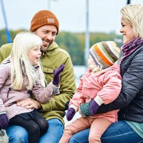 En mand og en dame sidder med hvert sit barn på skødet og i baggrunden er en havn