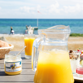 Juice, honning og andre lækre morgenmadsprodukter i forgrunden,. Strand, hav og blå himmel i baggrunden.